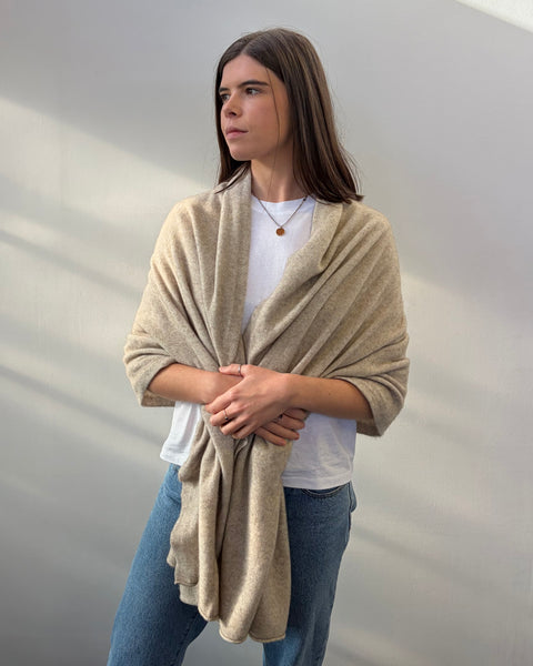 Woman wearing a beige shawl over a white shirt and blue jeans against a plain background