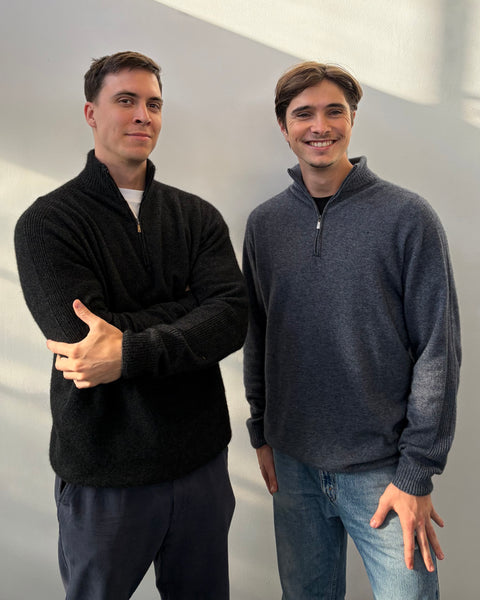 Two men standing side by side wearing dark grey and blue sweaters against a plain background