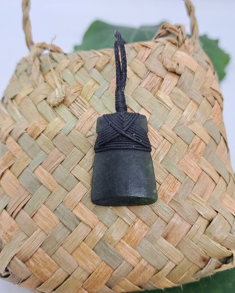 A dark carved greenstone necklace with a black cord wrapped around its upper section. Displayed again against a woven flax bag.