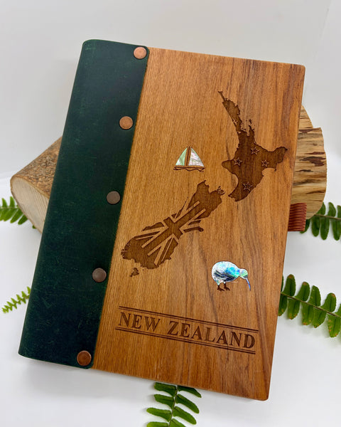 Wooden book with laser-cut map of New Zealand and 'New Zealand' engraving, surrounded by green leaves on a white background.