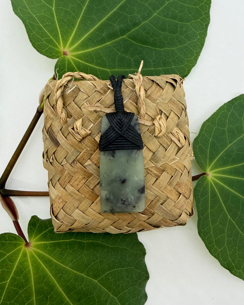 Greenstone pendant on a woven flax bag with a white background and green leaves
