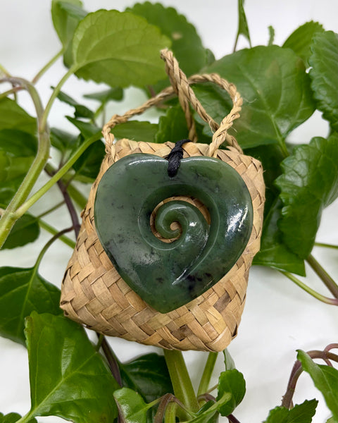 Heart-shaped greenstone pendant with a spiral in a woven basket against a leafy background