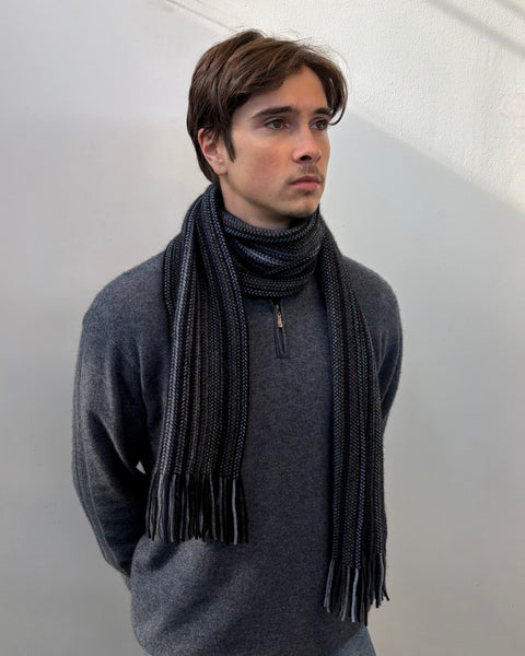 Man wearing a dark grey and navy knitted scarf and sweater against a plain background