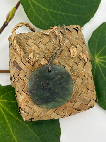 Greenstone pendant on a woven flax basket on a white background with green leaves