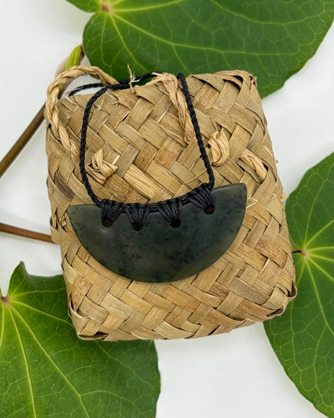 Crescent-shaped greenstone pendant with a bound black cord on a woven flax bag and green leaves on a white background