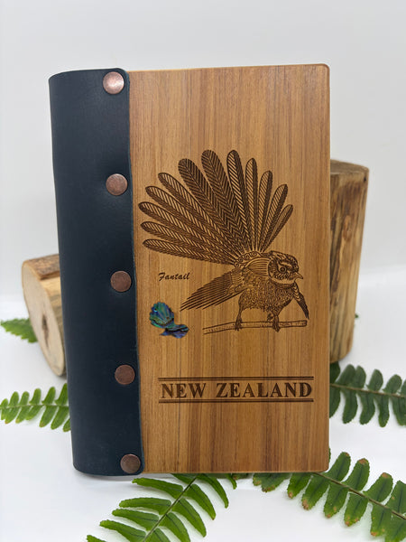 Wooden notebook with fantail illustration and 'New Zealand' text, navy leather cover spine, surrounded by green leaves.