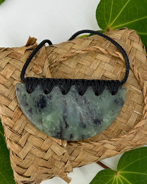 Greenstone pendant with a black cord on a woven flax basket on green leaves with a white background