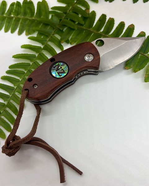 Folding knife with wooden handle and paua shell inlay on a white background with green leaves.