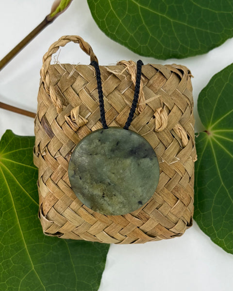 Greenstone pendant on a woven flax bag on a white background with green leaves
