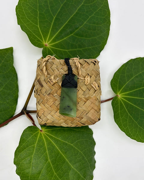 Greenstone pendant on a woven flax bag with a white background with green leaves