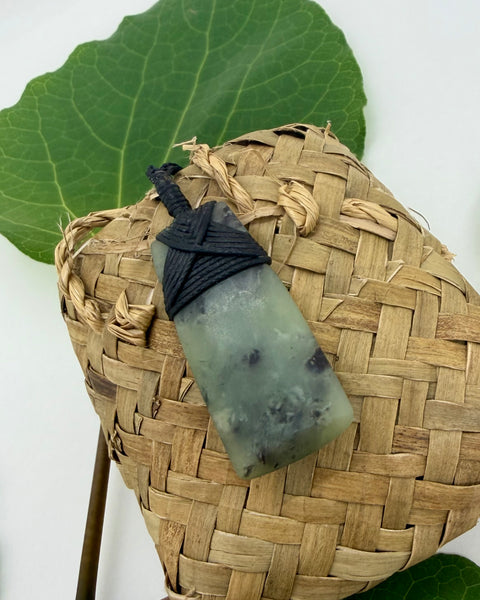 Greenstone pendant on black cord on a woven basket with green leaves in the background