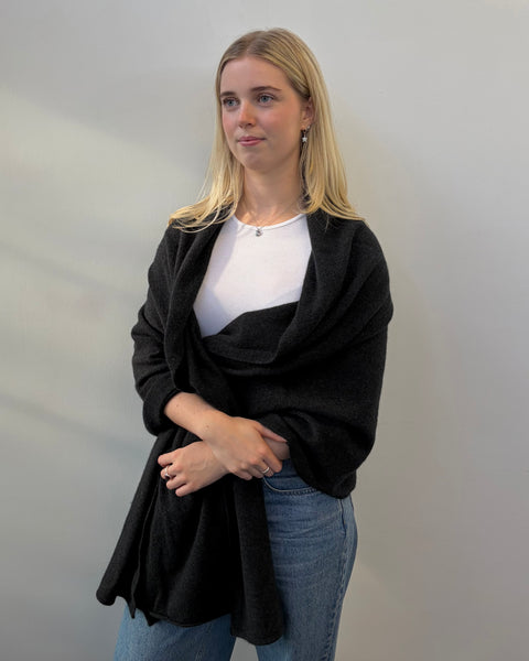 Woman wearing a dark grey shawl over a white shirt and blue jeans against a plain background