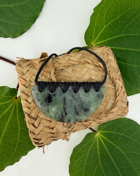 Crescent-shaped greenstone pendant with a black cord on a woven flax bag on free leaves with a white background