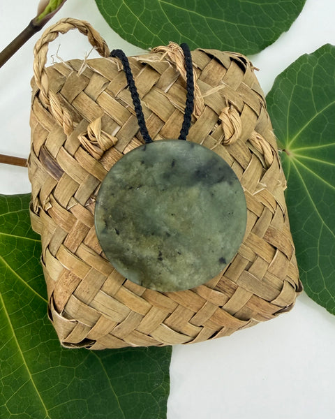 Greenstone pendant on a black cord laying on a woven flax bag with a white background and green leaves