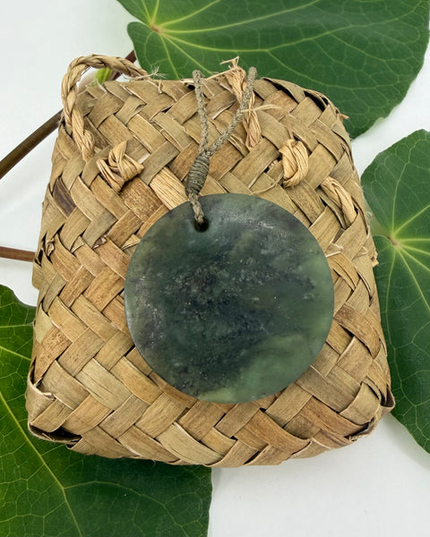 Greenstone pendant on a woven flax basket on a white background with green leaves.