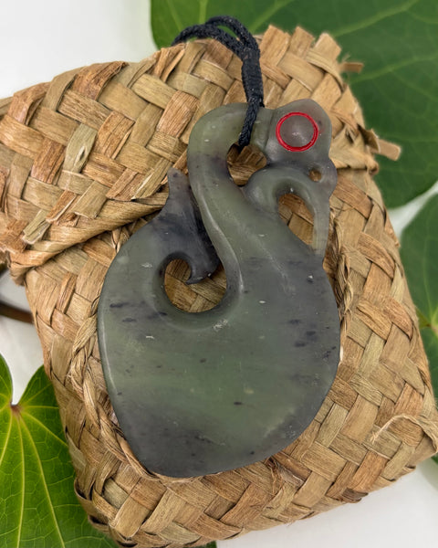 Greenstone manaia pendant on a woven flax bag on a white background with green leaves.