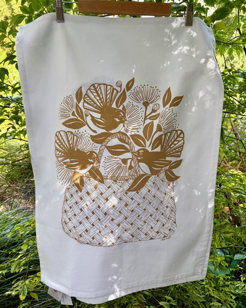 White tea towel with a gold graphic of Fantails and pohutukawa flowers over a woven flax bag.