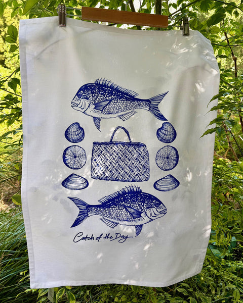 White tea towel with a blue graphic of two snapper and shellfish around a woven flax bag with the phrase 'Catch of the Day'.