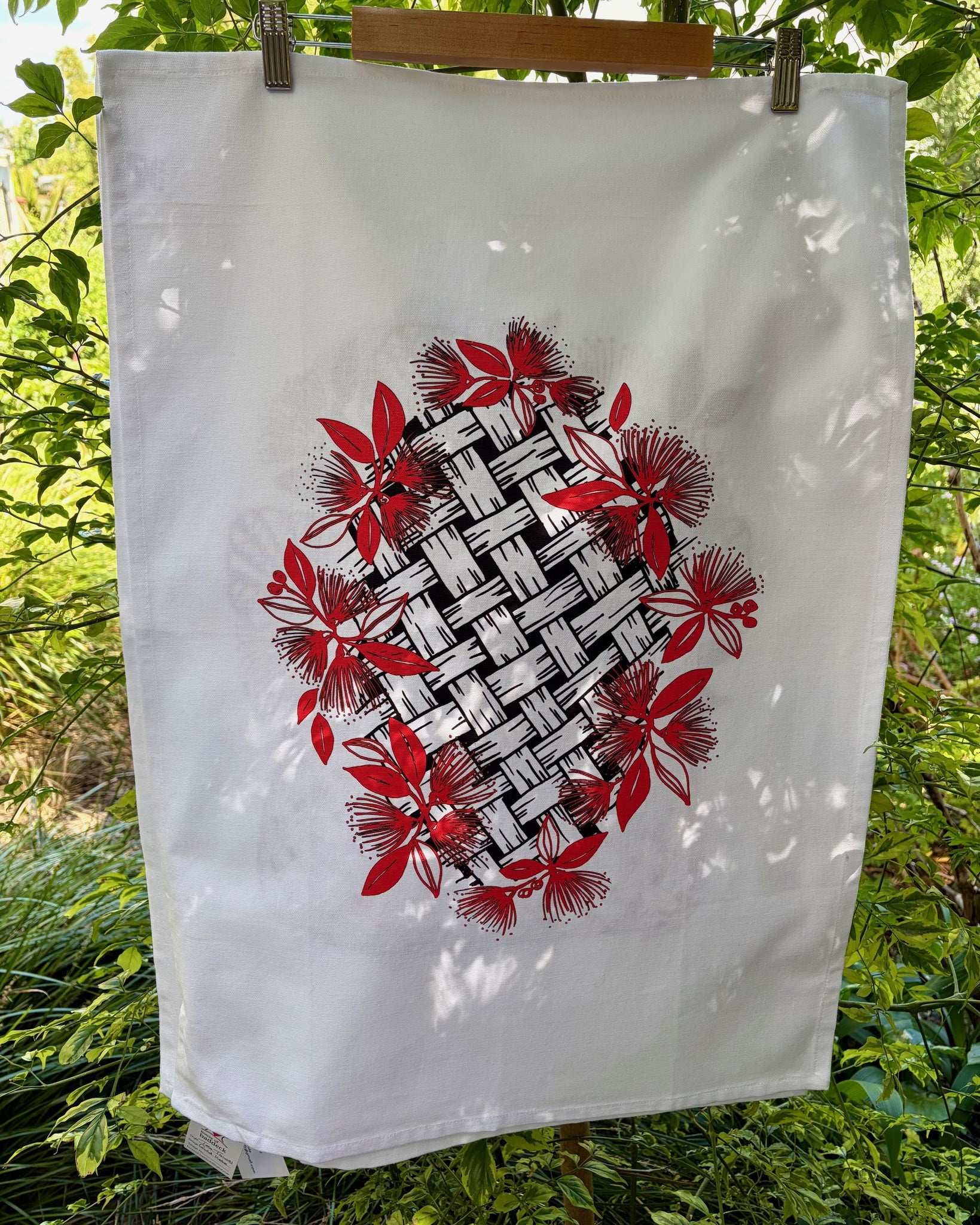 White tea towel with black woven flax pattern surrounded by red pohutukawa flowers.