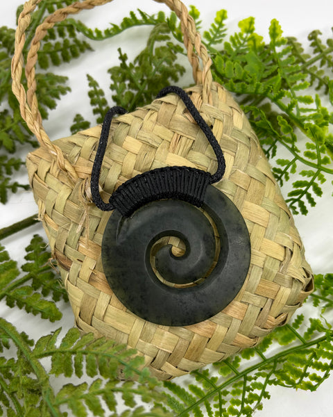 A carved greenstone pendant with a spiral design and a bound black cord sitting on a woven flax bag amongst green leaves