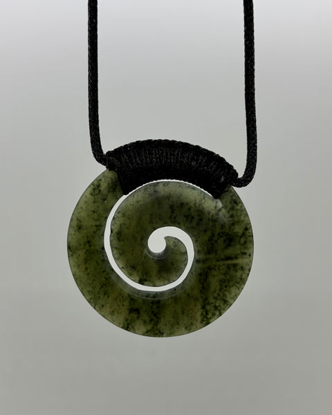 A greenstone pendant necklace with a carved spiral cutout design, suspended from a black woven cord on a neutral grey background.