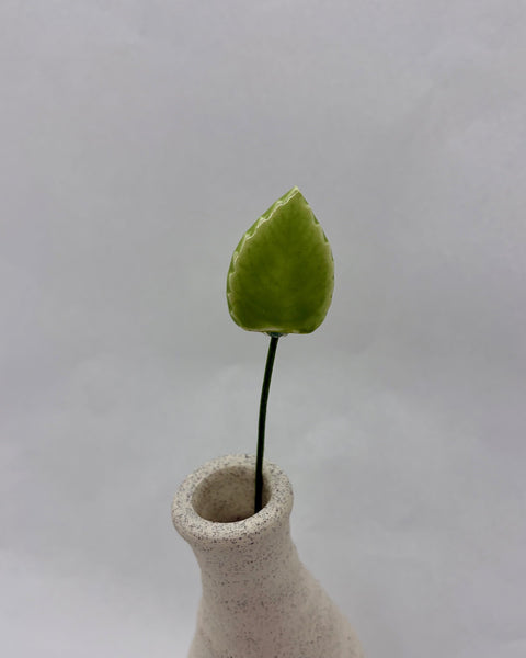 Green ceramic leaf with a thin green stem displayed in a white speckled vase.
