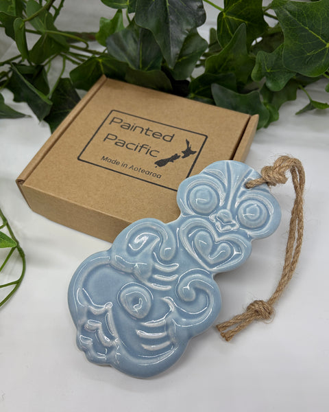 A light blue ceramic Tiki ornament with a jute hanging string, displayed next to its brown cardboard gift box labeled 'Painted Pacific' on a white surface with green leaves in the background.
