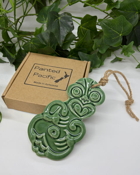 A mid green ceramic Tiki ornament with a jute hanging string, displayed next to its brown cardboard gift box labeled 'Painted Pacific' on a white surface with green leaves in the background.