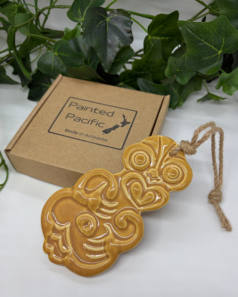 An ochre ceramic Tiki ornament with a jute hanging string, displayed next to its brown cardboard gift box labeled 'Painted Pacific' on a white surface with green leaves in the background.