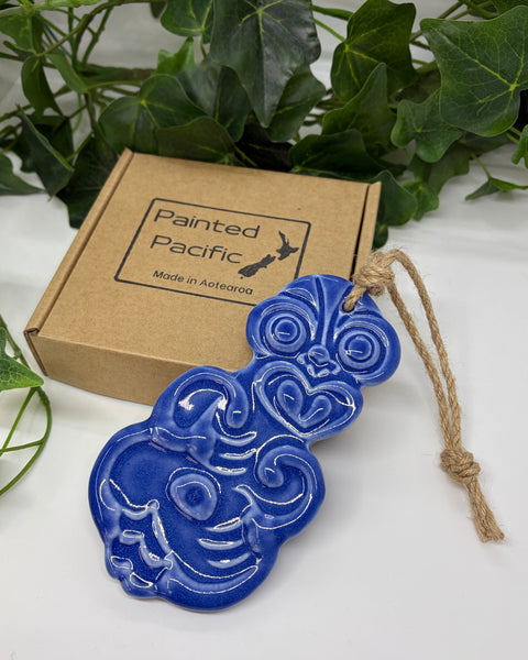 A purple ceramic Tiki ornament with a jute hanging string, displayed next to its brown cardboard gift box labeled 'Painted Pacific' on a white surface with green leaves in the background.