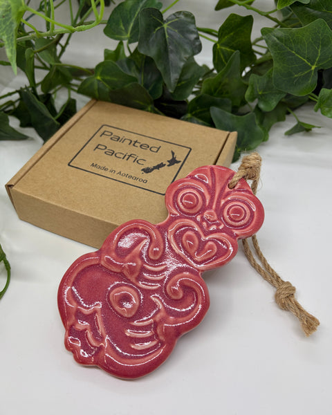 A soft red ceramic Tiki ornament with a jute hanging string, displayed next to its brown cardboard gift box labeled 'Painted Pacific' on a white surface with green leaves in the background.