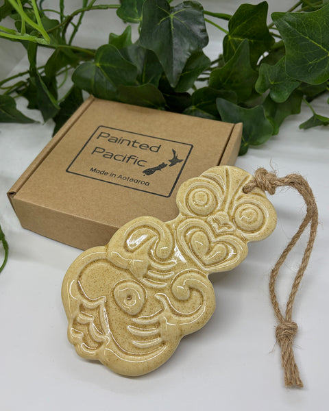 A sand coloured ceramic Tiki ornament with a jute hanging string, displayed next to its brown cardboard gift box labeled 'Painted Pacific' on a white surface with green leaves in the background.