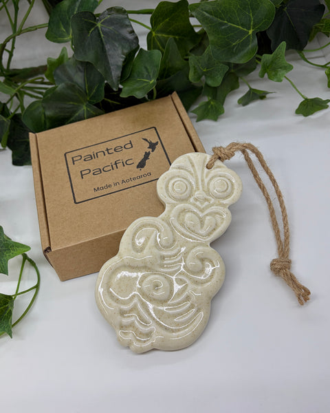 A sand white coloured ceramic Tiki ornament with a jute hanging string, displayed next to its brown cardboard gift box labeled 'Painted Pacific' on a white surface with green leaves in the background.