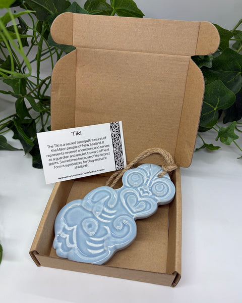 A light blue ceramic Tiki with a jute  hanging cord presented in an open brown cardboard gift box with a product card, set against a background of green houseplants.