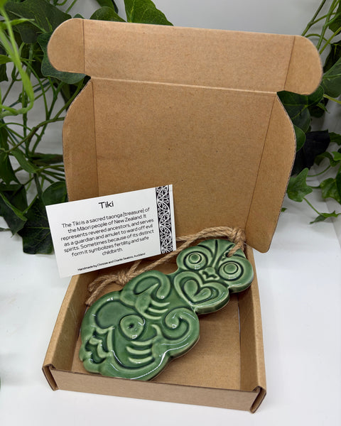A soft green ceramic Tiki with a jute  hanging cord presented in an open brown cardboard gift box with a product card, set against a background of green houseplants.