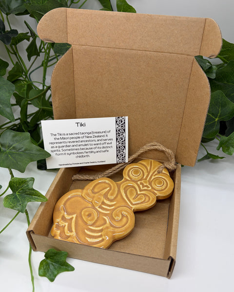 A tan ceramic Tiki with a jute  hanging cord presented in an open brown cardboard gift box with a product card, set against a background of green houseplants.