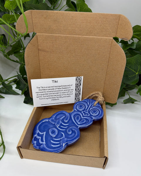 A purple ceramic Tiki with a jute  hanging cord presented in an open brown cardboard gift box with a product card, set against a background of green houseplants.