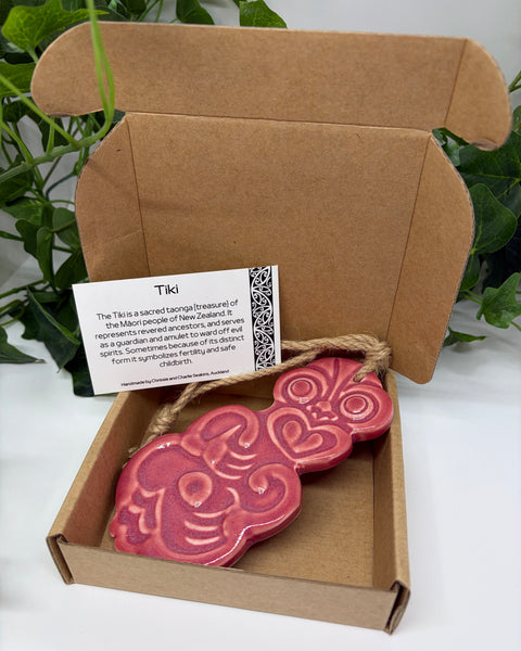 A light red ceramic Tiki with a jute  hanging cord presented in an open brown cardboard gift box with a product card, set against a background of green houseplants.
