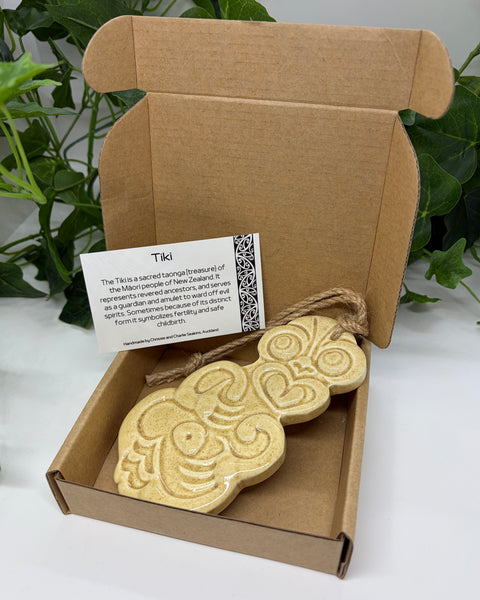 A sand coloured ceramic Tiki with a jute  hanging cord presented in an open brown cardboard gift box with a product card, set against a background of green houseplants.