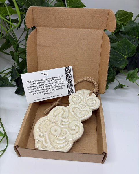 A sand white ceramic Tiki with a jute  hanging cord presented in an open brown cardboard gift box with a product card, set against a background of green houseplants.
