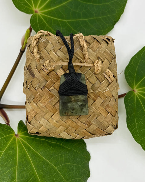 Greenstone pendant with a black cord on a woven flax bag with a white background and green leaves