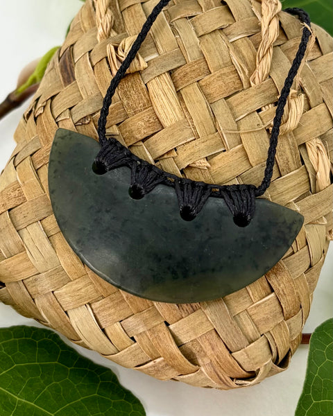 Crescent-shaped greenstone pendant with a black woven cord on a woven flax bag on a white background with green leaves
