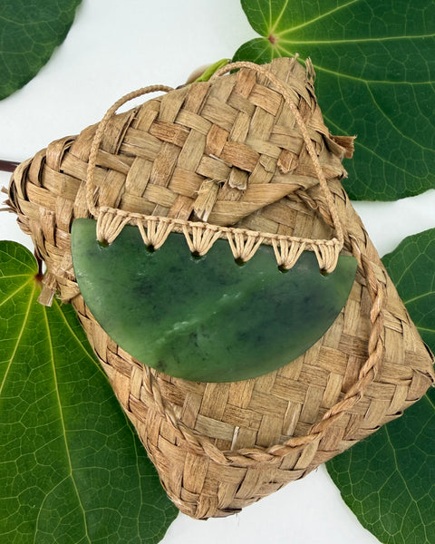 Greenstone pendant with a natural coloured cord on a woven flax basket and a leafy background