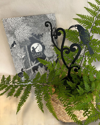 A black steel garden sculpture of a NZ native Huia Bird perched upon unfurling fern fronds, displayed in a pot plant and  with a hand-designed gift box