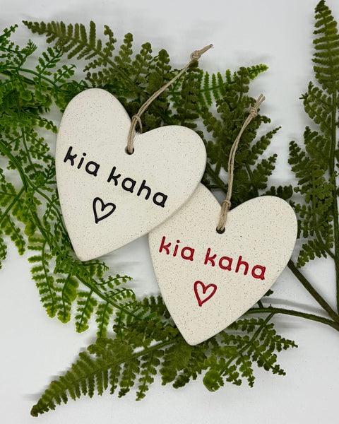 Two heart-shaped ceramic ornaments with natural speckled off-white glaze, hanging from twine strings. One ornament is printed with 'kia kaha' in black with a black heart, and the other is printed with 'kia kaha' in red with a red heart.