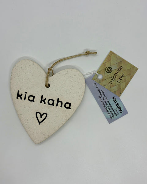 A heart-shaped ceramic ornament engraved with 'kia kaha' and a small heart, hanging from a twine string with a brand tag and product tag attached, on a plain white background.
