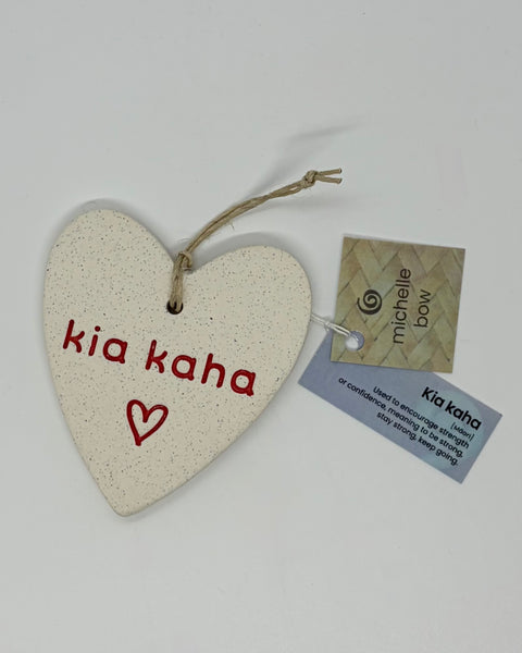 A cream coloured, heart-shaped speckled ceramic ornament with 'kia kaha' and a small heart in red, hanging from a twine loop. A paper tag attached to it reads 'kia kaha' with the English definition and shows the name 'michelle bow'.