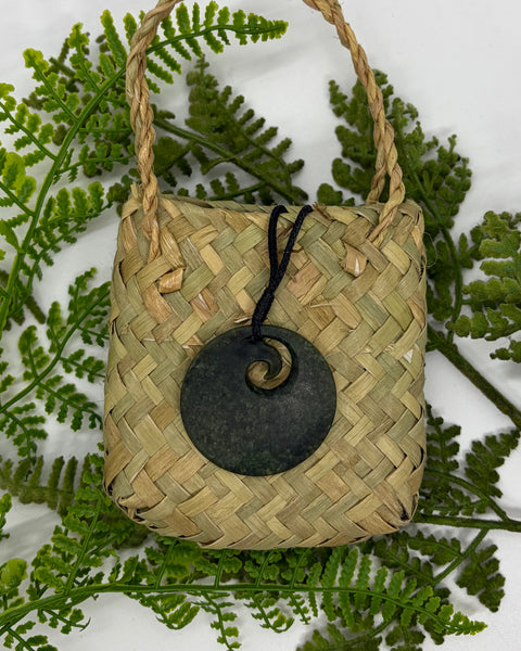A round greenstone pendant with a small spiral cutout and a woven black cord sitting on a flax bag amongst green fern fronds