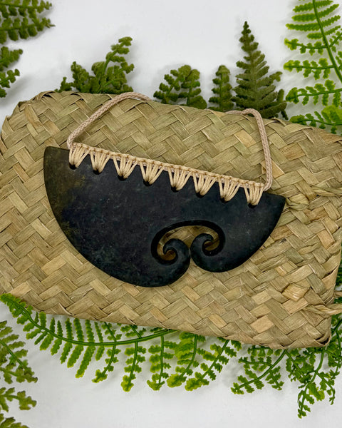 A carved greenstone pendant with a spiral cutout design and bound with a beige cord, sitting on a woven flax bag amongst green fern leaves.