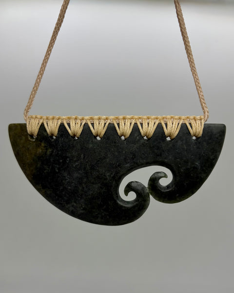A traditional Māori greenstone pendant with a carved spiral motif, suspended from a braided tan cord against a neutral grey background.